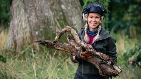 A woman wearing protective head gear carries dead wood in an area of woodland. There is a big tree trunk behind her and long grass.