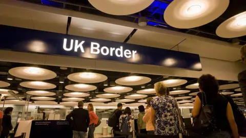 UK airport passport control. There is a blue overhead sign with UK border written on it in white. People are queuing to get their passports checked.