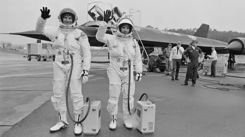 Getty Images A black and white image showing American airmen Maj William C Machorek, Jr, and Capt Harold B Adams in their spacesuit-like flightsuits, waving, with their American USAAF reconnaissance aircraft, the Lockheed SR-71 Blackbird, in the background at RAF Mildenhall in Suffolk, England, 12th September 1974. 