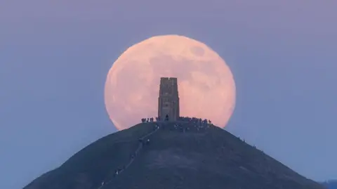Weather Watchers/Infocus A large supermoon behind Glastonbury Tor. There are crowds of people up on the hill. The sky is a light blue