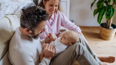 A dad sat on the floor with baby on his lap. Mum looks on from his side.