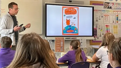 Tom stands in front of a smart board with children in purple school jumpers watching him describe how to draw the characters from his book.