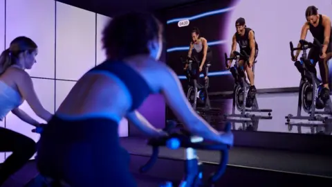 Wiltshire Council Two women on stationary exercise bikes pointing at a screen of people also on exercise bikes