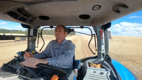 a picture of joe inside the cab of a blue tractor. In the background we can see the fields and skyline.
