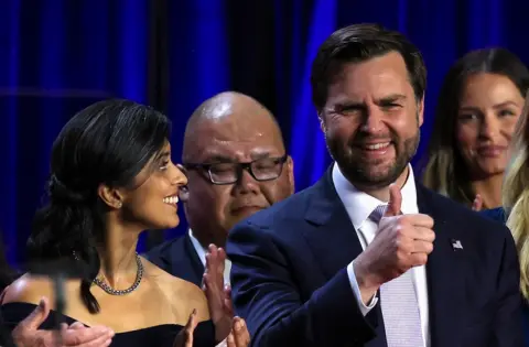 Getty Images Usha Vance in a sleeveless dress looking at JD Vance who is giving a thumbs-up sign