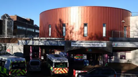 The outside of the Royal Surrey County Hospital which has a large circular top part of the building clad in red metal. There is a sign with the hospital name on it and outside there are ambulances parked and a low rise building seen in the background.