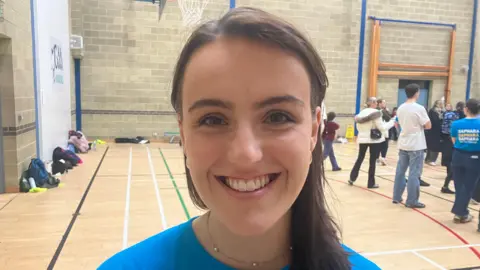 Leah is wearing a blue t-shirt, she has long brown hair and a small chain around her neck. She is standing in a busy sports hall with parents and children behind her. 