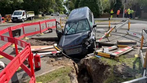 Andrew Turner/BBC Another angle of the Land Rover crashed in the gas mains trench. Works in high-vis clothing stand nearby in discussion.