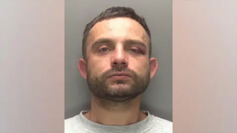 A police head and shoulders shot of a man with short brown hair looking into the camera with a serious expression. He has brown stubble and a moustache and is wearing a grey top. One of his eyes appears drooped and bruised.