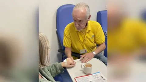 An older man with slicked back grey hair holds a short-haired woman's hand in a hospital setting. He wears a yellow NHS T shirt, a lanyard, and sits in a blue chair. The woman is in a mossy green fleece and has a bandage on her hand.