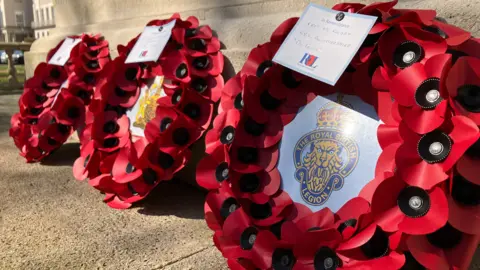 Three poppy wreaths, each with Royal British Legion symbols in the middle, lay at the bottom of a war memorial in a town centre on a sunny day. They have handwritten "In Remembrance" notes on them.