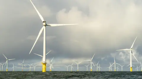 PA Media Large offshore wind turbines rise from the sea on tall yellow foundations, stretching into the distance across the water. Their white blades rotate against a sky filled with thick, layered clouds, while choppy waves move across the surface below.