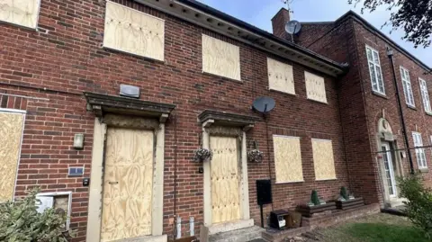 A terraced row of brick-built homes, with two whose doors and windows that were destroyed in the fire having now been boarded-up. To the right a third property apparently looks unaffected.