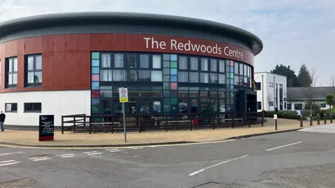 The Redwoods Centre. Built in the round, the upper part of the building is covered in red wooden panels, with the lower part in white render. The front of the building, under a large sign reading "The Redwoods Centre" features lots of glass windows