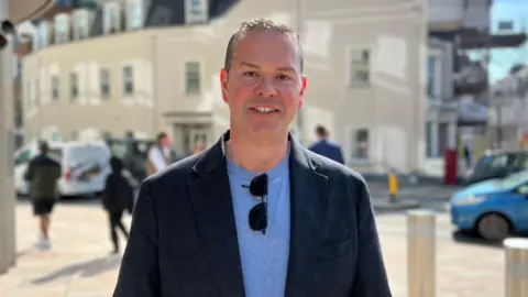 Marcus Calvani wearing a dark blazer over a light shirt with sunglasses hanging from the collar. In the background, several people walk along the pavement, and a light‑coloured building with scaffolding on its upper section is visible. 