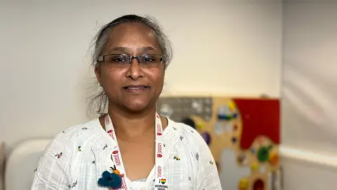 BBC Rikha Wilmot is wearing a white shirt with a white lanyard around her neck. In the background there is a children's play area.