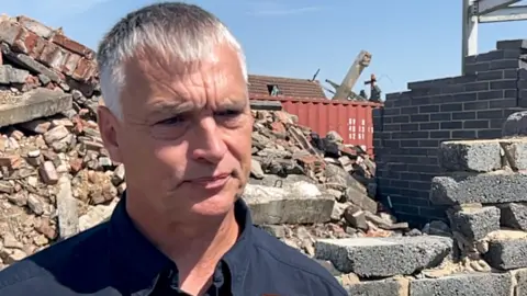 Burchnall stands in front of part of a breeze block wall with lots of rubble behind it. He wears a dark blue top.