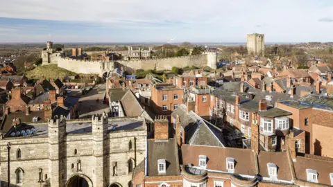 A bird's eye view of the city of Lincoln. Rows of houses are in the foreground, with a castle wall on a hill in the distance from Lincoln Castle.