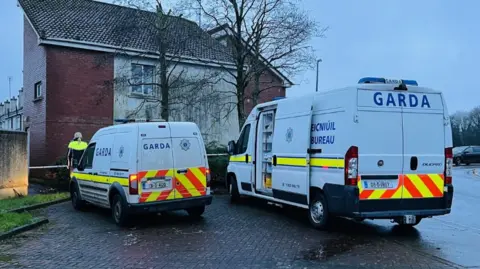 RTÉ Two white Garda vans are parked outside a domestic property behind Gardaí tape. An officer is seen standing in front of one of the vans.