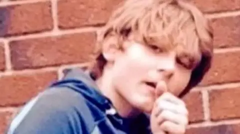 The late Daniel Gee Jamieson, who was murdered aged 16 in 2018. He is photographed with long brown hair and is wearing a blue hooded tracksuit top. Daniel has his hand near his mouth while doing a "thumbs up" gesture. He is outside standing in front of a brick wall.