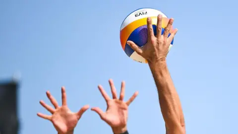 A close-up of a person hitting a volleyball and two hands trying to defend it. The sky is blue.