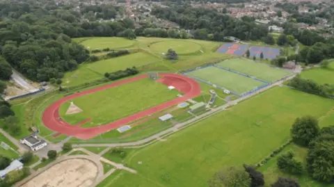 Department for Levelling Up, Housing and Communities An aerial view of the sports centre, showing an athletics track, circular cricket pitches, artificial football pitches and tennis courts