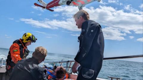 A coastguard winchman and two people in dive suits stand next to a man laying down on a boat which is at sea. Above their heads is a red and white Coastguard Rescue helicopter.