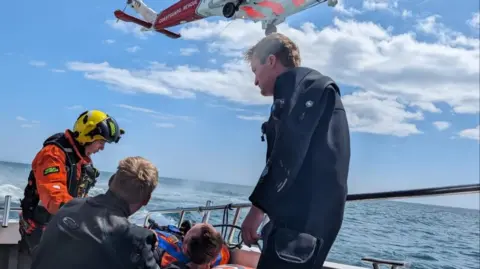 A coastguard winchman and two people in dive suits stand next to a man laying down on a boat which is at sea. Above their heads is a red and white Coastguard Rescue helicopter.