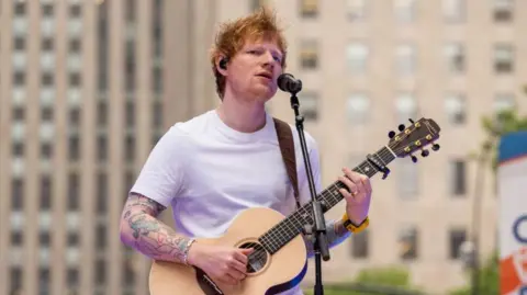 Getty Images Ed Sheeran playing an acoustic guitar at an outside gig in 2023. He is wearing jeans and a white t-shirt.