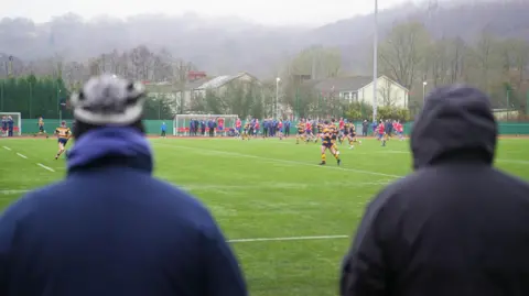 A picture of Aberdare's first team playing rugby against Penallta RFC. The picture has been taken behind two men who are watching the game.