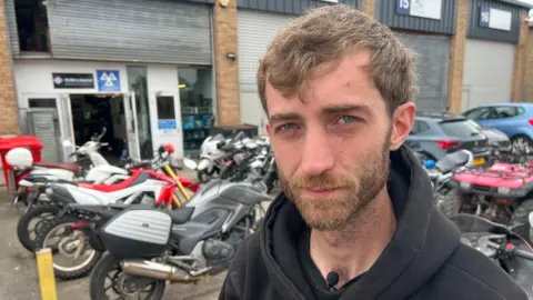 Jamie, who has short cropped brown hair and a beard. He is wearing a black hoodie and standing in front of a collection of motorbikes