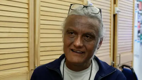 A man with grey hair and glasses, wearing a white top underneath a dark blue jacket, standing in front of light brown wardrobes. 