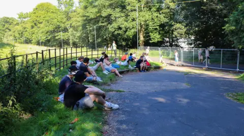 Emma Gibbs A number of Oasis fans in T-shirts, shorts and trainers sit in the shade of a wooded area alongside a path, which has been cordoned off with metal wire fencing, separating fans with Oasis tickets from those without