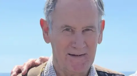 Head and shoulders photo of Richard Parker. He has grey hair and is wearing a check shirt and brown jacket. He is looking into the camera with a slight smile. Someone is standing next to him out of shot but they have their arm around him and their hand is on his shoulder.