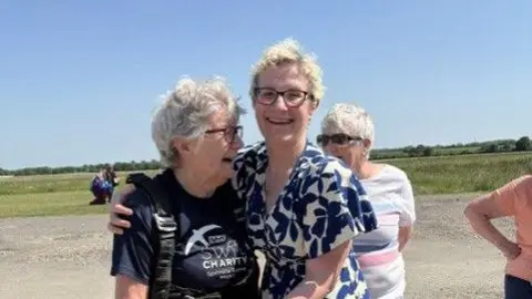 Avril Abraham is wearing a parachute harness stands on an airfield, being hugged by Sarah Allen in a patterned dress. Avril is wearing a dark T-shirt with a charity logo. In the background, a few other people stand on the gravel area, with open fields and a clear blue sky behind them.