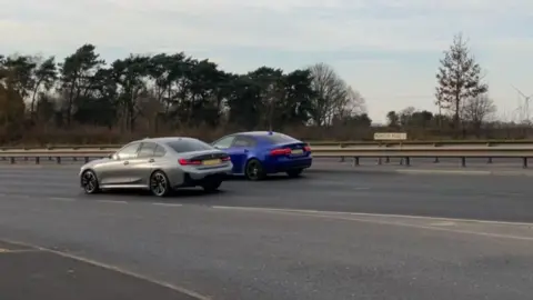 Two BMW cars on the dual carriageway at dusk, not during a race.