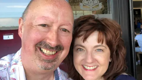 Mark Cowgill A smiling middle-aged couple. He has a close-shaved head and goatee beard. She has shoulder length auburn hair. There is a 'Bantams' sign behind them, showing they are at a football stadium.
