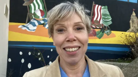 BBC/Lara King A woman with short blonde hair smiles as she stands in front of a brightly coloured mural depicting striped deckchairs, striped towels and a sandcastle on a beach. She is wearing pearl stud earrings, a beige jacket and a blue top.