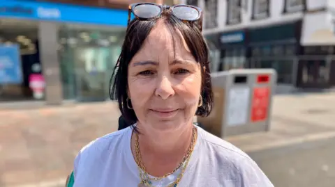 Julie is standing in Cardiff city centre, wearing a white t-shirt, with her glasses on her head, looking at the camera. She has short brown hair, and has a gold chain necklace and gold hoop earrings. 
