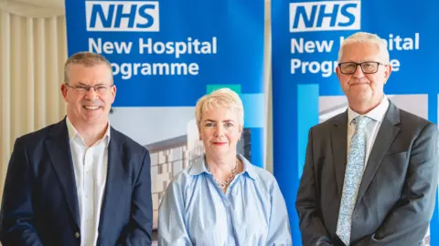 NHS England A photo of three people, showing Joe Harrison Karin Smyth and Pat Boyle. All standing by two signs, that are blue and white. They are all looking at the camera. Joe has on glasses, a white shirt and blue jacket, Karin, a blue shirt, short blonde hair and a necklace, and Pat, has short grey hair, glasses and is wearing a white shirt, tie and grey jacket. 