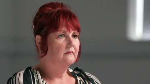 A grab of Linda Larkin from her TV interview. She is loking slightly off camera as she is interviewed with a blurred out background. She has red hair and a black and white dress.