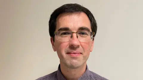 Dr Gavin McCloskey smiling at the camera as he stands in front of a beige wall. He has short black hair and is wearing thin framed glasses. He is also wearing a purple and white checked shirt.