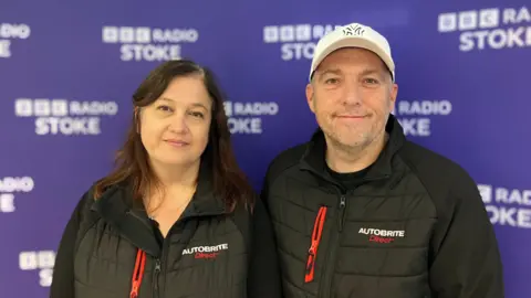 Jill and Mark Moss, wearing black Autobrite Direct branded gilets, are stood against a purple BBC Radio Stoke branded backdrop.