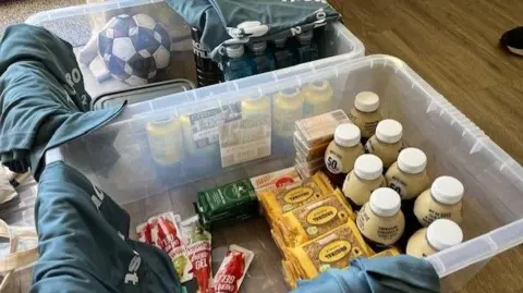 BBC/James Phillips Two plastic baskets with drinks and snacks and grey sports tops. There is also a football.