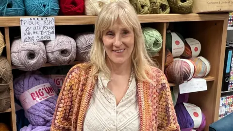 Susie Goate - who runs crafts and fabric shop SewSusieBloom - says she wants the newly elected councillors to build on projects, across Castleford, that are designed to bring the community together, is pictured in her shop. She is standing in front of a rack of huge balls of wool, all different colours. She is smiling at the camera, has shoulder-length blonde hair and is wearing a white blouse and crocheted cardigan.