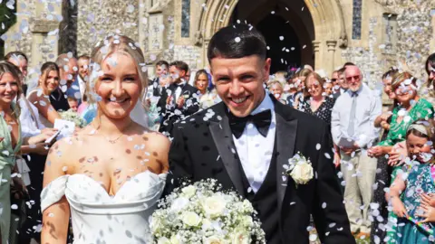 Contributed A wedding photo of a couple as they emerge from church, showered in confetti. She is to the left and wearing a shoulderless white wedding dress. She has blonde hair. He is wearing a black bow tie and tuxedo, and has dark short hair. Both are smiling, as smiling guests look on from behind. 