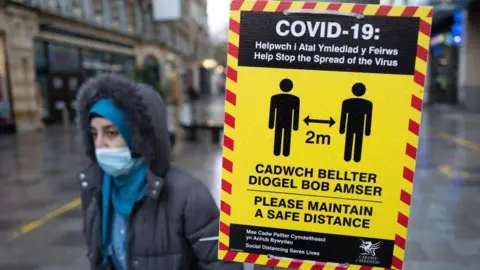 A bilingual Covid-19 sign in a high street encouraging two metre social distancing during the pandemic. A pedestrian wearing a face mask can be seen beside the sign. The pedestrianised street is shiny and wet from rain.