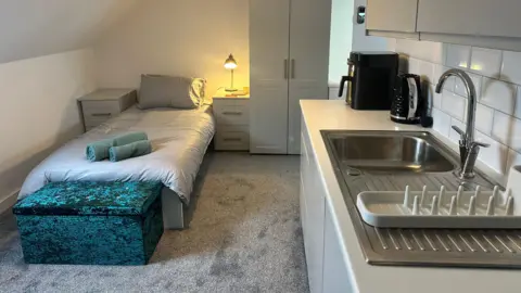 A modern-looking living space with a single bed, a wardrobe, chests of drawers and a kitchenette. The walls are white, while the decor is co-ordinated with a grey carpet, furniture and bedding, and a blue ottoman and towels. A bedside lamp glows a warm yellow colour. The kitchenette has light grey cupboards and surfaces, an aluminium basin and a black kettle and coffee machine.