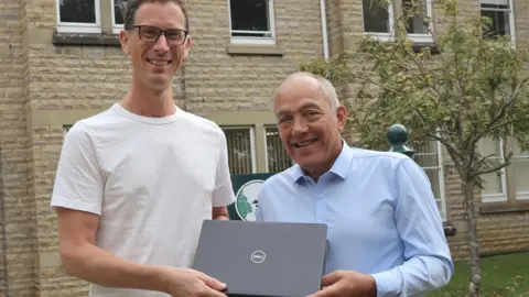 Ben Tuppin from Getting Oxfordshire Online (L) and councillor Andy Graham (R) smiling at the camera while holding a DELL laptop. There is a building and a tree behind them.