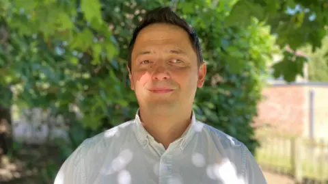 Man in a white shirt standing in a field in front of a tree smiles at the camera